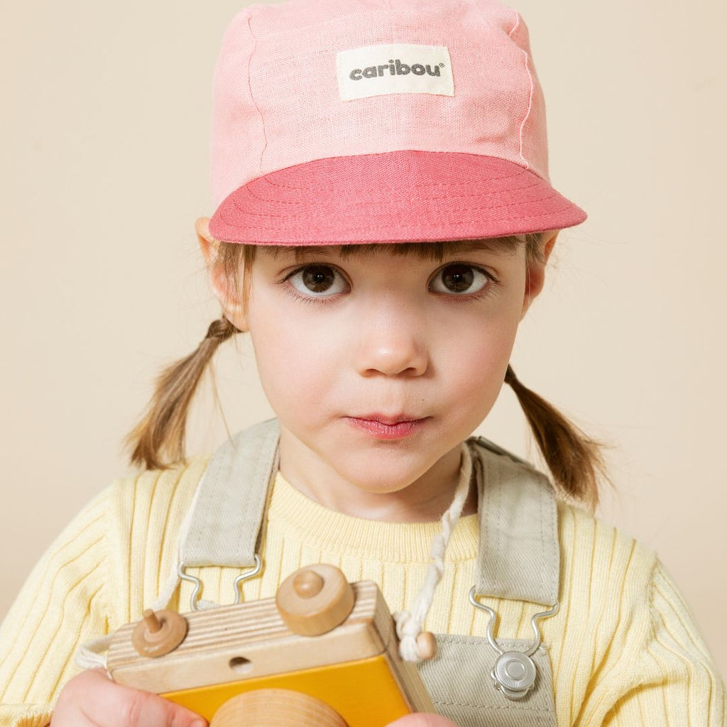 Une photo d'une petite fille radieuse portant la Casquette en Lin Duo Rose de Caribou. Elle tient un appareil photo en bois dans ses mains et semble impatiente de capturer des souvenirs.