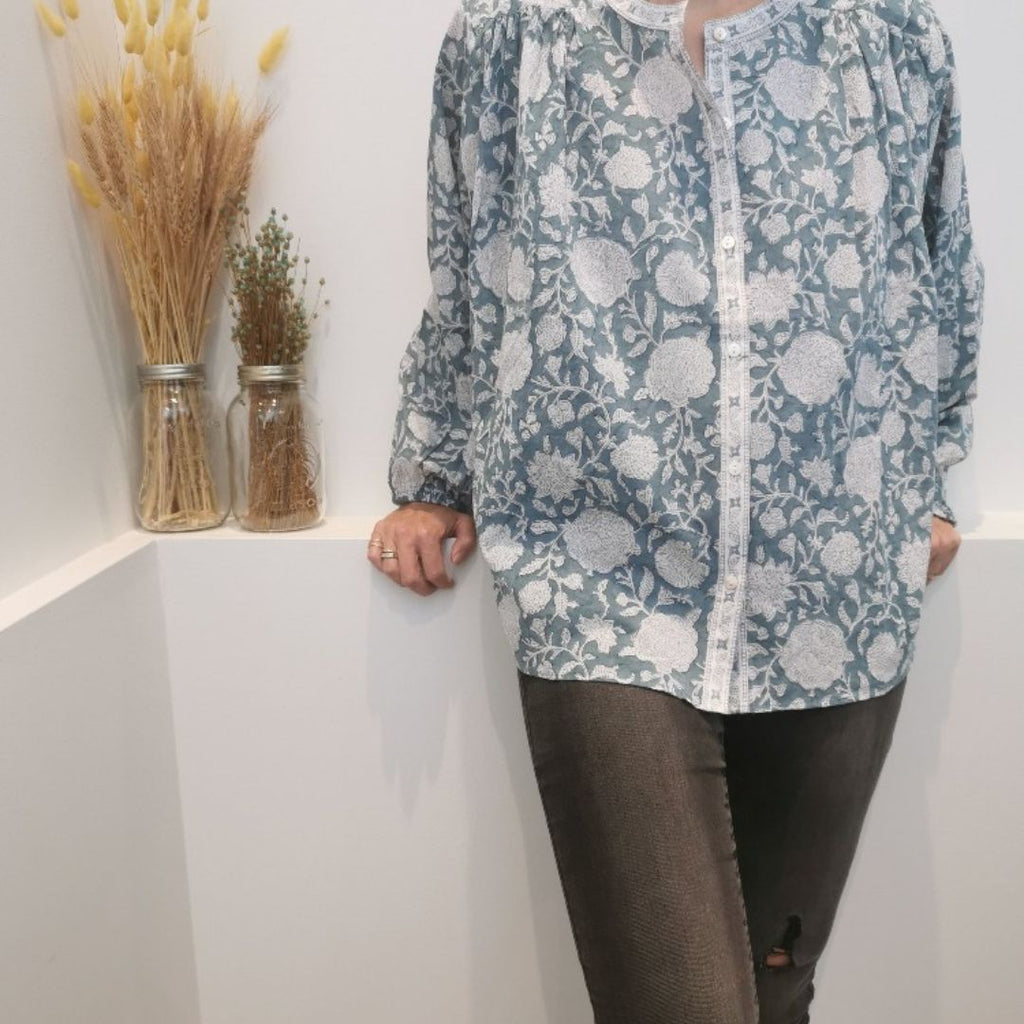 Une élégante blouse Bonheur du jour Paris "Oriana" de couleur Tupia bleue, portée par une femme. La blouse, d'une forme fluide et aérienne, est fabriquée en mousseline de coton et possède des manches courtes bouffantes et un col montant.