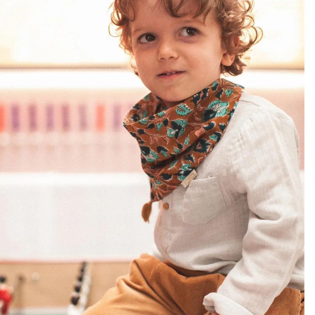 Un adorable petit garçon souriant jouant et vêtu d'une chemise Bonheur du jour Paris "Elliott" en double gaze de coton bleu clair. La chemise, d'une coupe droite et décontractée, est réhaussée de petits boutons blancs et d'un col classique avec son foulard imprimé au blockprint