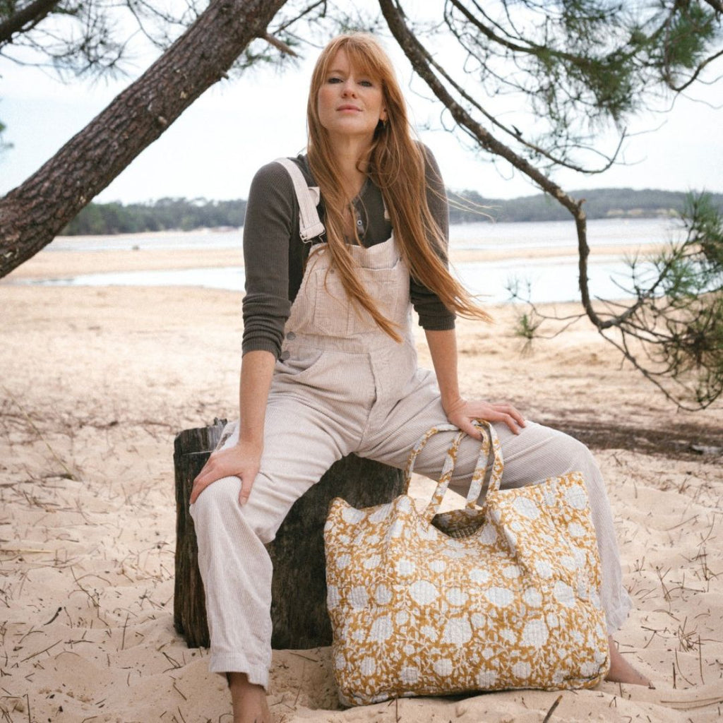 Une femme rousse souriante porte un sac shopper Tupia Absinthe de Bonheur du Jour Paris, La femme porte une tenue décontractée et bohème, les pieds dans le sable