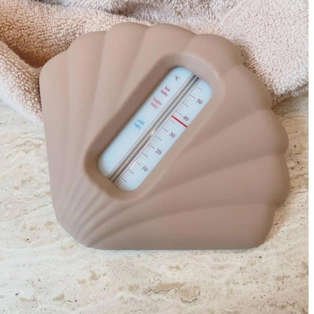 Un thermomètre de bain en silicone de chez Konges Slojd, adorable en forme de coquillage rose poudré, posé sur le bord d'une baignoire remplie d'eau.